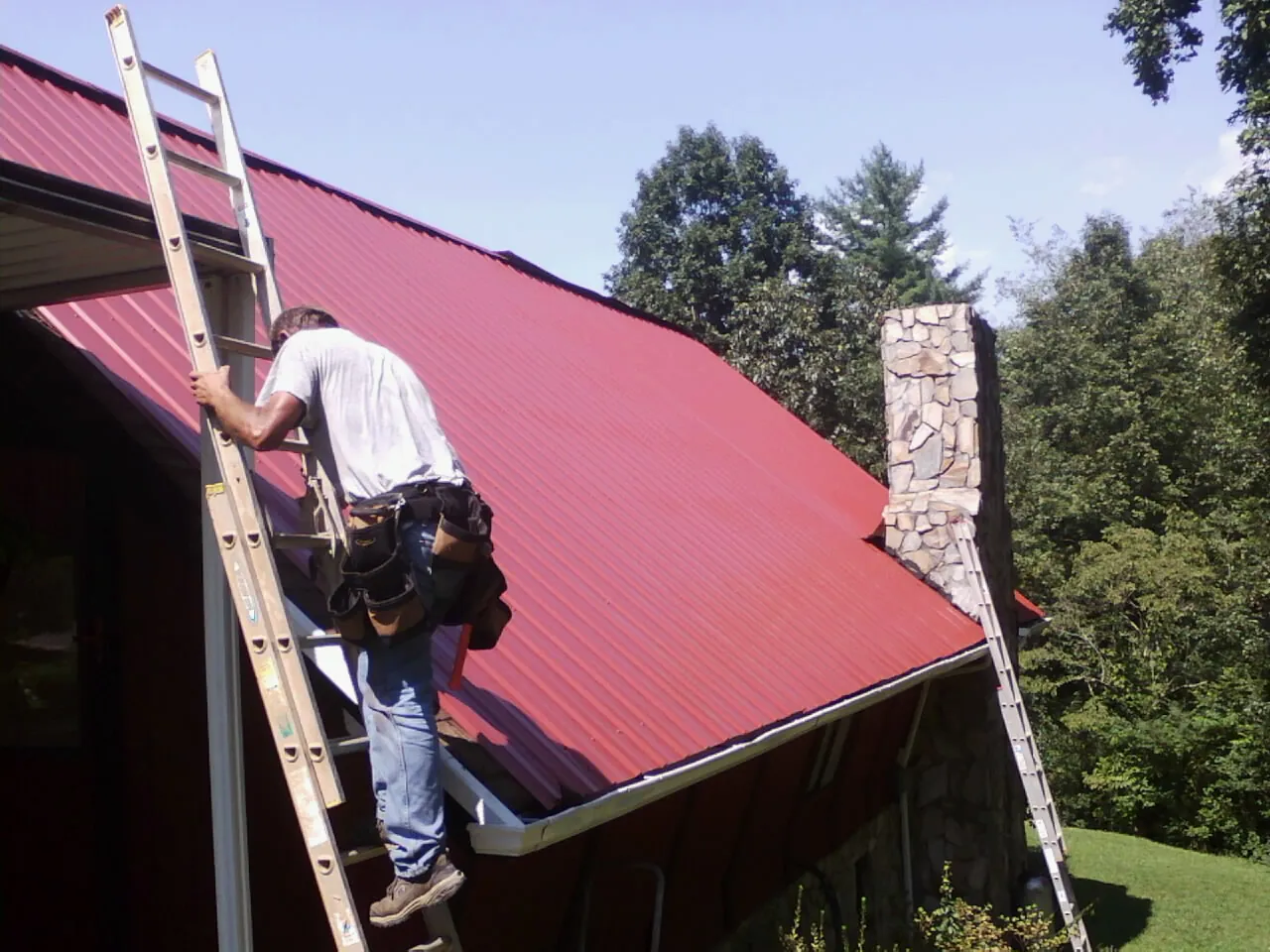 Roofer on a ladder beside a red metal roof and stone chimney — Soffit & Fascia Repair by Roofing Brothers in Jackson Springs